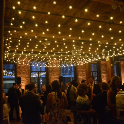 A canopy of String Lights suspended above the dance floor with warm white up-lights placed at the base of brick columns between windows for a wedding at The Dumbo Loft located in Brooklyn New York
