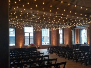 A canopy of String lights suspended between center columns over ceremony are with warm white up-lights placed at the base of each column between windows for a wedding at The Dumbo Loft located in Brooklyn New York