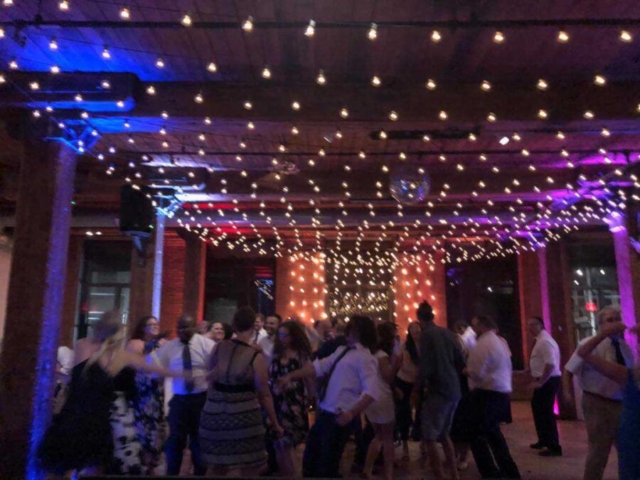 A canopy of String lighting hanging betweeen the center columns along with string lights hanging vertically behind ceremony at The Dumob Loft