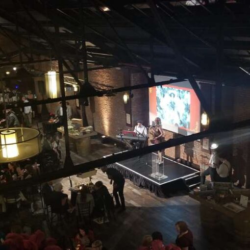 A Projector suspended overhead from a Truss with a screen mounted on the wall and a spotlight with additional AV equipment and stage was provided for the Louie Awards hosted at 26 Bridge in Brooklyn, NY.