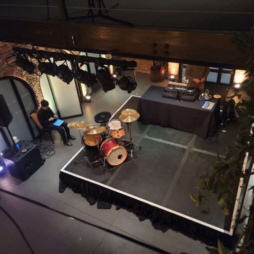 A 12ft x 12ft stage in the main room under the mezzanine level near the entrance doors with dimmable spotlights suspended overhead at The Foundry.