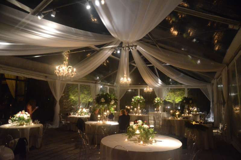 Decorative white drapes with Crystal Chandeliers and Pin-Spots focused on the floral centerpieces on each table under a clear top tent in the courtyard at The Foundry.