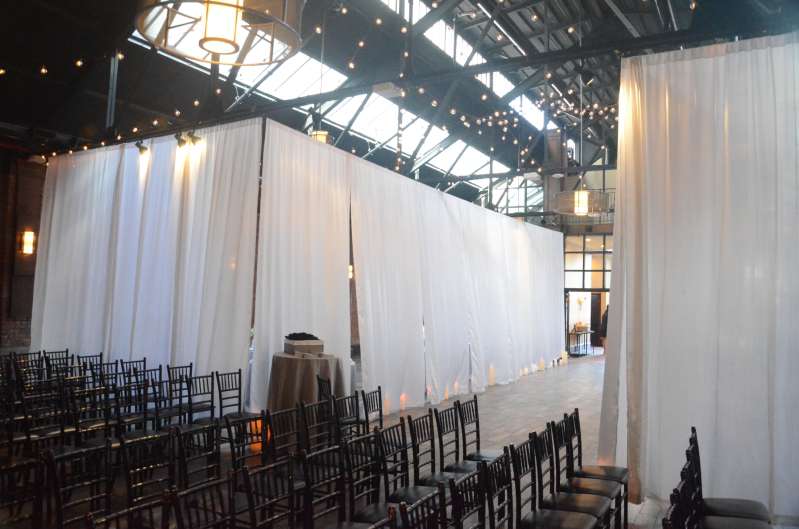 White Sheer curtains are hanging on the main floor at 26 Bridge. String lights are hanging in two circular patterns between the 1st beam and the 4th beam in the main room at 26 Bridge.