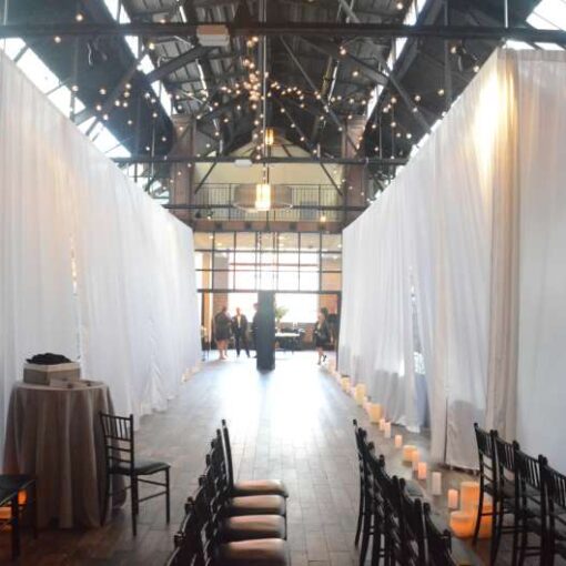 White Sheer curtains are hanging on the main floor at 26 Bridge. String lights are hanging in two circular patterns between the 1st beam and the 4th beam in the main room at 26 Bridge.