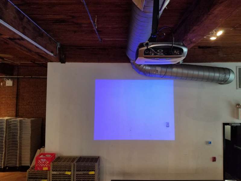 A Projector hanging overhead and displaying a video image on the White Wall adjacent to the corner emergency exit at The Dumbo Loft.
