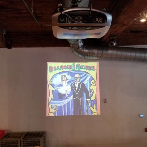 A Projector hanging overhead and displaying a video image on the White Wall adjacent to the corner emergency exit at The Dumbo Loft.