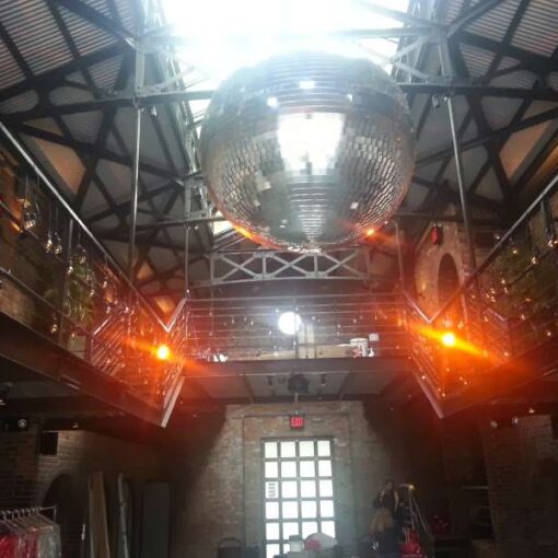 A 40" Disco Ball hanging between the railing of the mezzanine level in the main room at The Foundry. String Lights draped along the railing of the mezzanine level.
