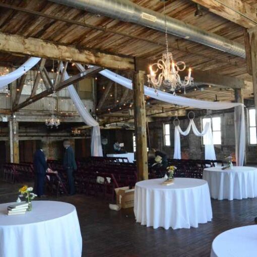 Sheer curtains hanging in an X-Shaped pattern under the high ceiling area of the main floor with String Lights and a chandelier.  White sheer drapes hanging in an M-Shaped pattern with three white rings for the wedding ceremony.