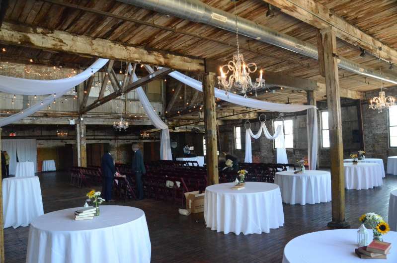 Sheer curtains hanging in an X-Shaped pattern under the high ceiling area of the main floor with String Lights and a chandelier.  White sheer drapes hanging in an M-Shaped pattern with three white rings for the wedding ceremony.