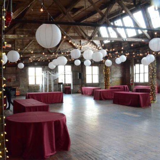 String lights are hanging in a circular pattern with white paper lanterns, and additional string lights are wrapped around the six center columns for a wedding in the main room at The Greenpoint Loft.