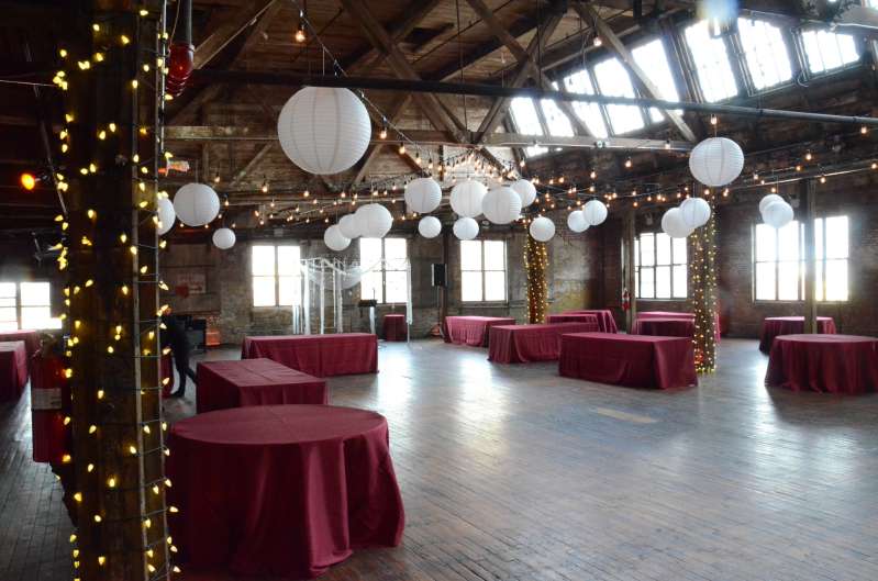 String lights are hanging in a circular pattern with white paper lanterns, and additional string lights are wrapped around the six center columns for a wedding in the main room at The Greenpoint Loft.