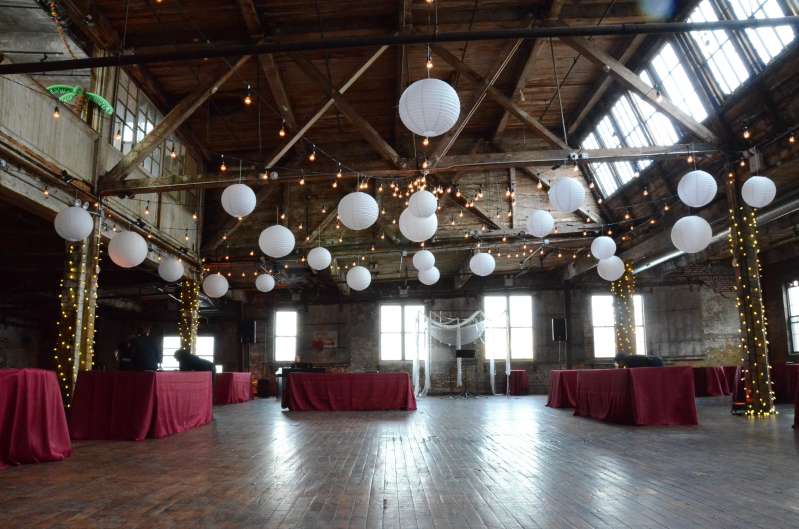 String lights are hanging in a circular pattern with white paper lanterns, and additional string lights are wrapped around the six center columns for a wedding in the main room at The Greenpoint Loft.