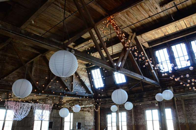 String Lights hanging with large swoops leading up to a center point above the ceiling in an hourglass shape pattern with White Paper Lanterns for a wedding at The Greenpoint Loft.
