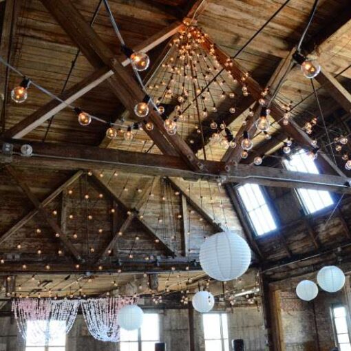 String Lights hanging with large swoops leading up to a center point above the ceiling in an hourglass shape pattern with White Paper Lanterns for a wedding at The Greenpoint Loft.