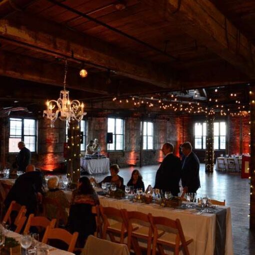 String lights are hanging in a zigzagging pattern with additional string lights wrapped around the six center columns and chandeliers for a wedding in the main room at The Greenpoint Loft.