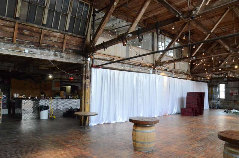 String Lights with S14 Bulbs are hanging under the high ceiling area between the six center columns on the main floor for a wedding at The Greenpoint Loft. Also, white Non-Sheer Curtains for partitioning the ceremony area.