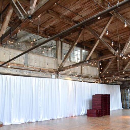 String Lights with S14 Bulbs are hanging under the high ceiling area between the six center columns on the main floor for a wedding at The Greenpoint Loft. Also, white Non-Sheer Curtains for partitioning the ceremony area.