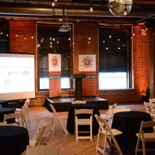 A  projector hanging overhead from a ceiling mount with screen at The Dumbo Loft. Also Podium and Stage for an fundraising event for the Brooklyn Workforce Innovations at the Dumbo Loft.  Also, string lights hanging over the center of the main room. Finally, amber up-lights around the perimeter of the room.
