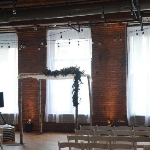 White sheer curtains hanging in each window for a wedding at The Dumbo Loft.  Also, string lights hanging over head between the center columns at The Dumbo Loft. Finally, Up-Lights setup at the base of each brick column between the windows.