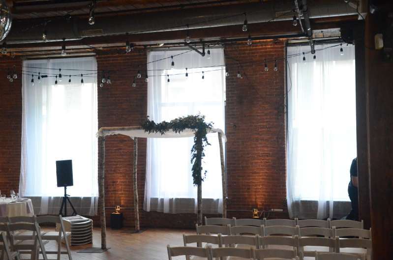 White sheer curtains hanging in each window for a wedding at The Dumbo Loft.  Also, string lights hanging over head between the center columns at The Dumbo Loft. Finally, Up-Lights setup at the base of each brick column between the windows.