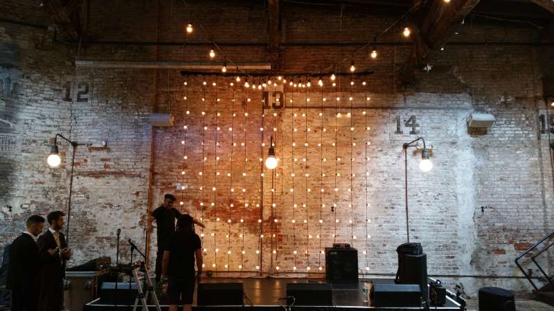 Vertical String Lights hanging against the wall as a backdrop behind the ceremony area for a wedding at The Houston Hall. Also setup a 18ft x 8ft stage to elevate the band.