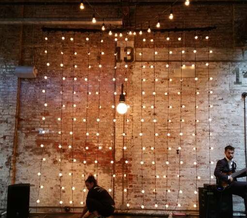 Vertical String Lights hanging against the wall as a backdrop behind the ceremony area for a wedding at The Houston Hall. Also setup a 18ft x 8ft stage to elevate the band.