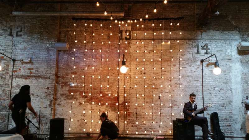 Vertical String Lights hanging against the wall as a backdrop behind the ceremony area for a wedding at The Houston Hall. Also setup a 18ft x 8ft stage to elevate the band.