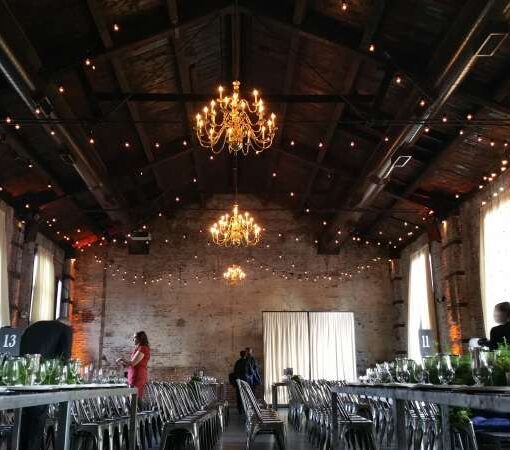String Lights hanging in a zigzagging pattern over the majority of The Green Building for a wedding. Amber Up-Lights are also set up along the perimeter walls of the main room at The Green Building for a wedding.