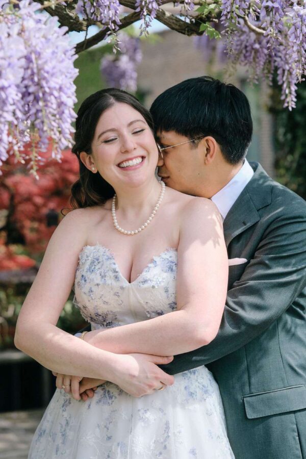 A joyful bride in a floral dress and pearls smiles as her partner hugs her from behind under blooming wisteria. Jocelyn & Andrew's wedding at The Foundry in Long Island City, NY.