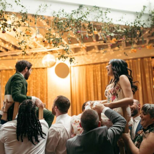 Sua & Adam are lifted by family and friends during the hora during their wedding reception in The Atrium at Rule of Thirds, with the shimmery light from a mirror ball visible in the background.
