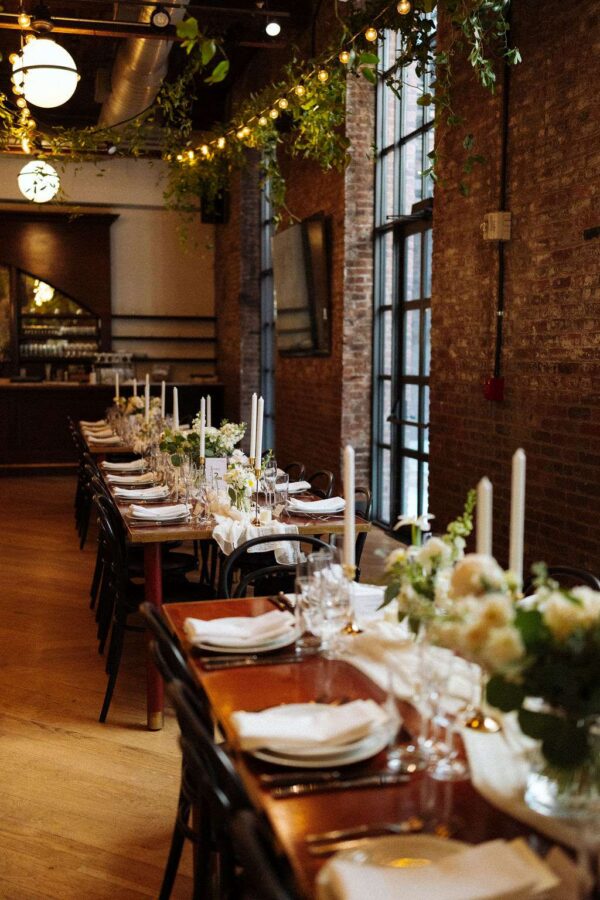This image displays lighting tips for creating an elegantly decorated reception space at The Wythe Hotel, ideal for a wedding, showcasing the room's rustic charm with green florals and warm, white string lights hanging overhead.