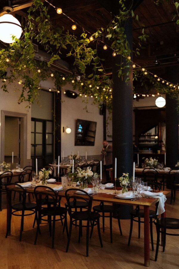 This image displays lighting tips for creating an elegantly decorated reception space at The Wythe Hotel, ideal for a wedding, showcasing the room's rustic charm with green florals and warm, white string lights hanging overhead.