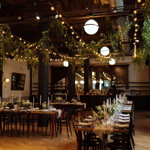 This image displays lighting tips for creating an elegantly decorated reception space at The Wythe Hotel, ideal for a wedding, showcasing the room's rustic charm with green florals and warm, white string lights hanging overhead.