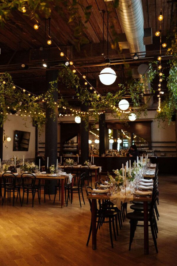 This image displays lighting tips for creating an elegantly decorated reception space at The Wythe Hotel, ideal for a wedding, showcasing the room's rustic charm with green florals and warm, white string lights hanging overhead.