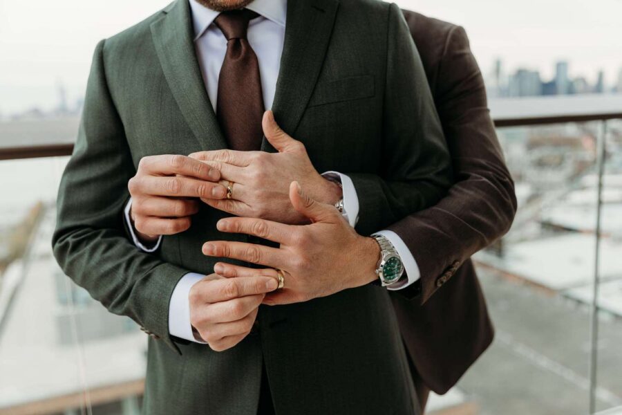 An intimate moment of affection, with a close-up of two formally dressed men sharing a tender embrace, highlighting their hands and wedding rings.