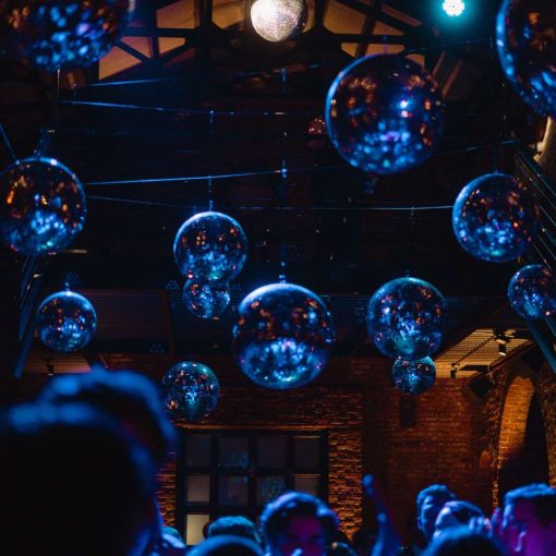 Guests are dancing in the main room under multiple mirror balls with a lavender color wash on the dance floor. Guests are dancing in the main room under multiple mirror balls with a lavender color wash on the dance floor.